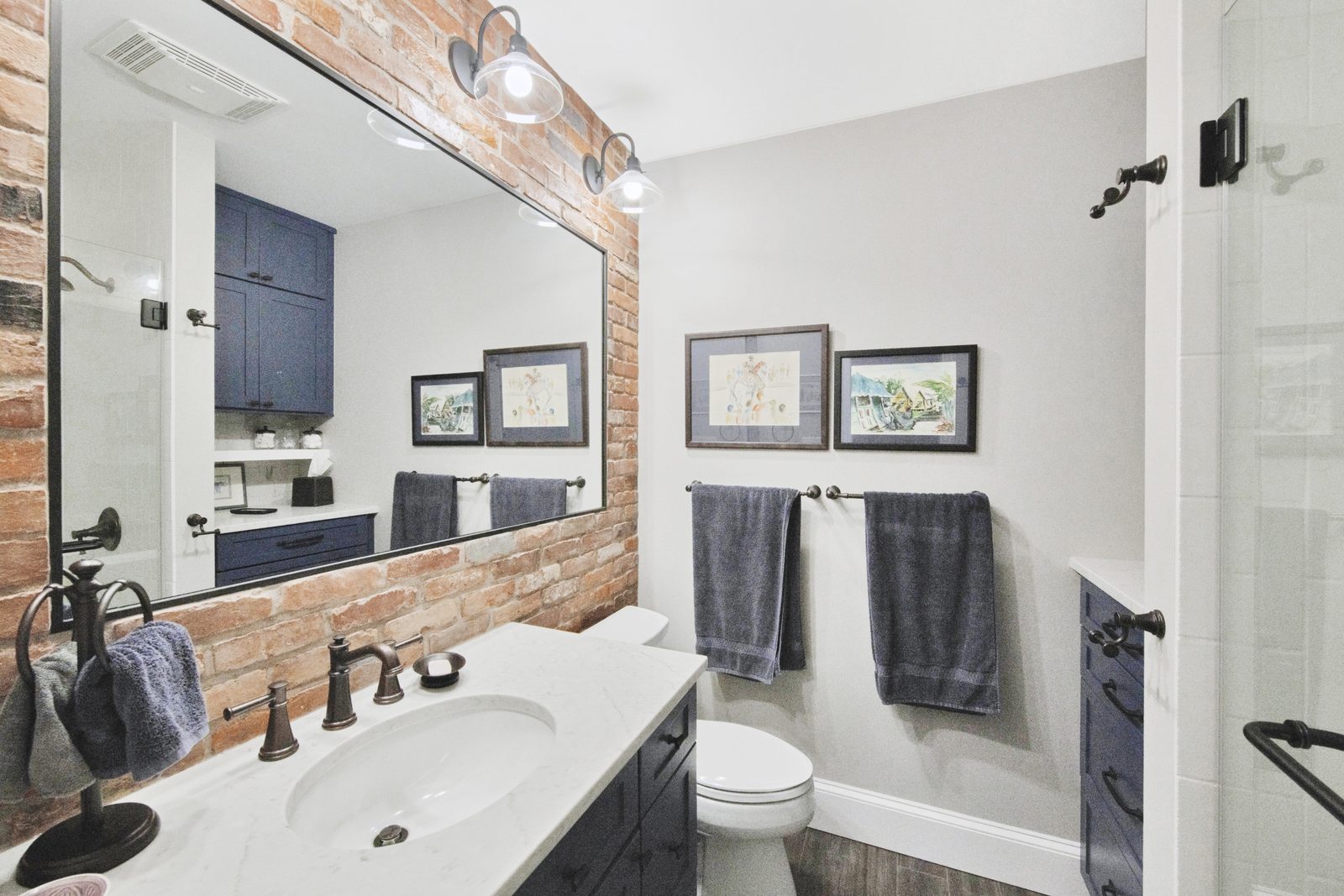 Bath — brick accent wall with framed mirror, navy cabinetry, walk-in shower with marble