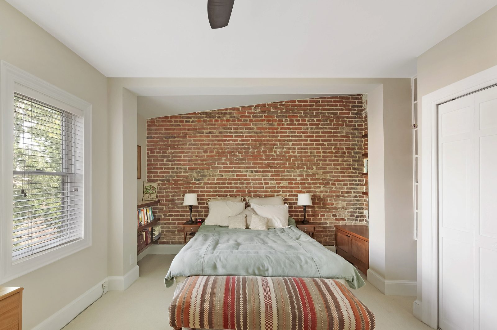 Bedroom — full-height exposed brick wall, ceiling fan, large window, striped ottoman bench